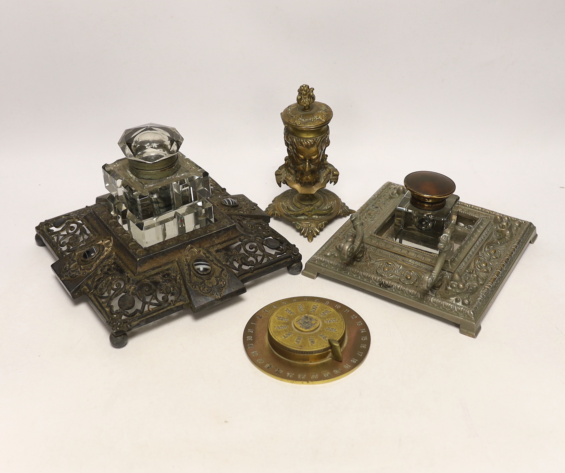 Three cast metal and cut glass ink stands, including a bronze inkwell in the form of the head of Bacus, and a brass calendar display from the Banca Commerciale Italiana, London Branch, tallest 13cm